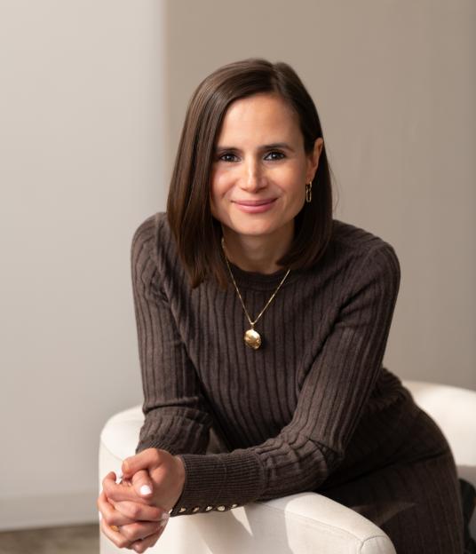 A woman with medium-length brown hair, wearing a brown ribbed sweater and a golden pendant necklace, smiles directly at the viewer.