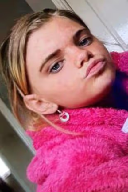 A young girl with blue eyes and blonde hair wears a fuzzy pink jacket and a circular earring.