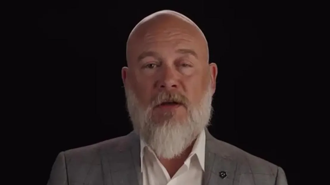 Bryan Seaver, bald with a white beard, wearing a plaid blazer and white shirt, speaks in front of a black background.