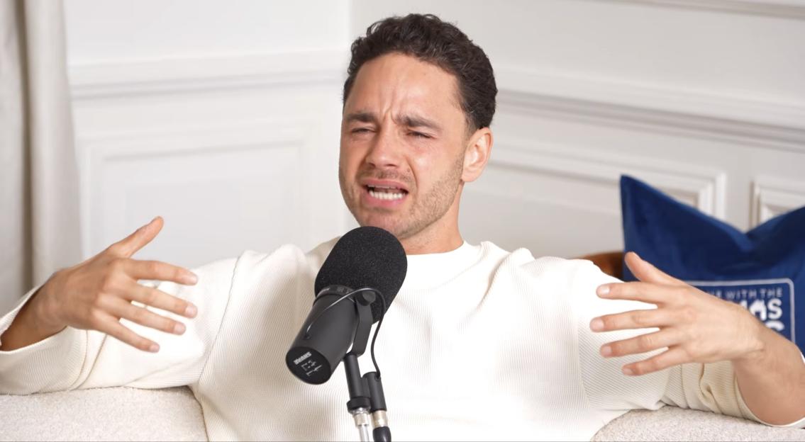 Adam Thomas speaking into a microphone, gesturing with his hands, and looking upset.