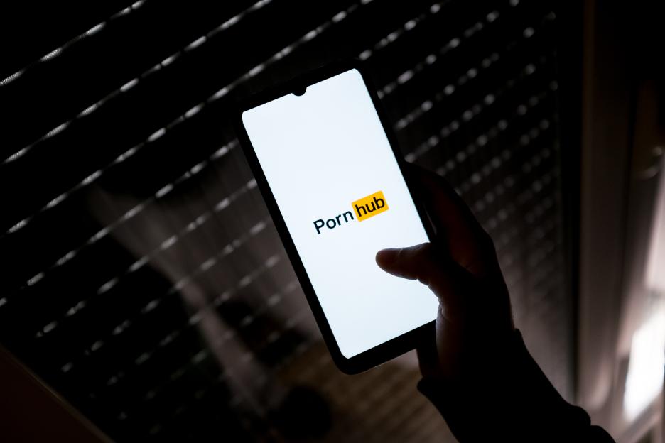 A smartphone displaying the Pornhub logo is held in a silhouetted hand against a dark, striped background.