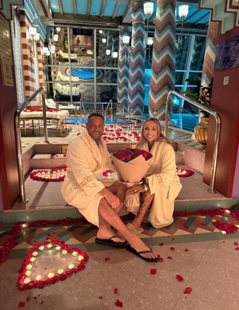 A couple in robes sits surrounded by rose petals and candles, holding a bouquet of red roses.