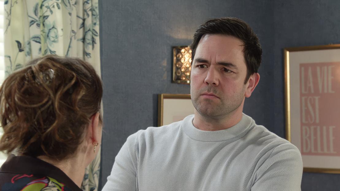 A man with dark hair in a light blue sweater looks with suspicion at a woman seen from behind.