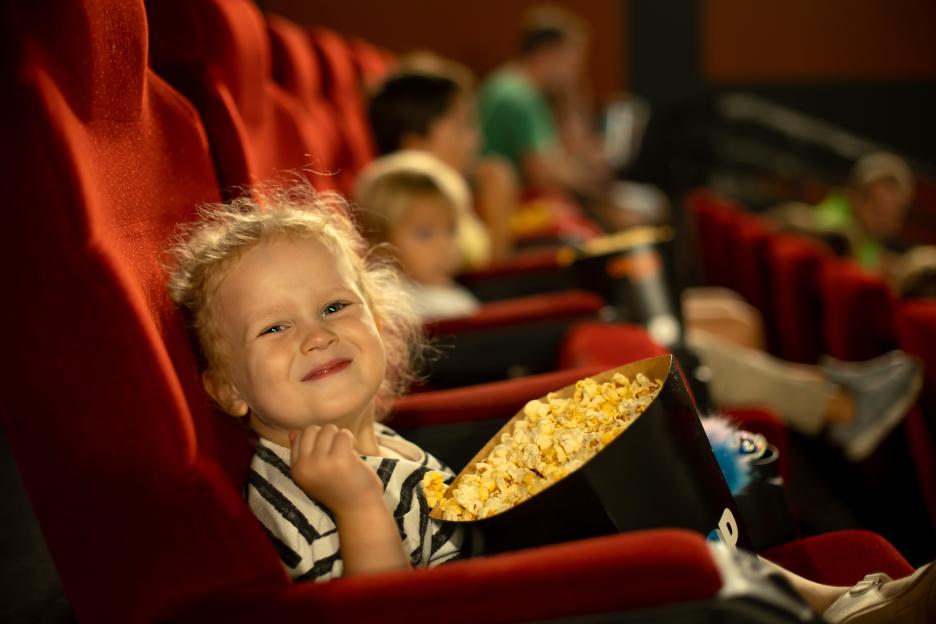 Cute child, curly girl, watching movie in a cinema, eating popcorn and enjoying