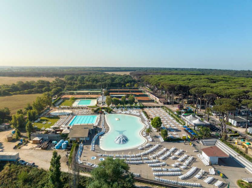 Aerial view of a resort with multiple swimming pools and tennis courts surrounded by fields and a large forest.