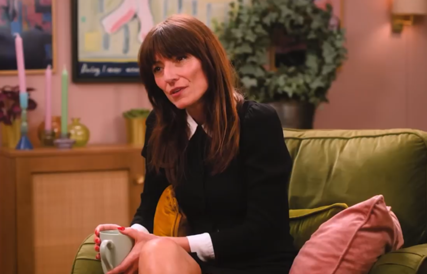 A woman in a black dress with a white collar, holding a light green mug, sits on a green couch.