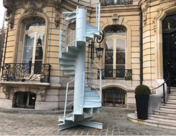 A 24-step spiral staircase, once part of the Eiffel Tower, stands on a cobbled street in front of an ornate building.