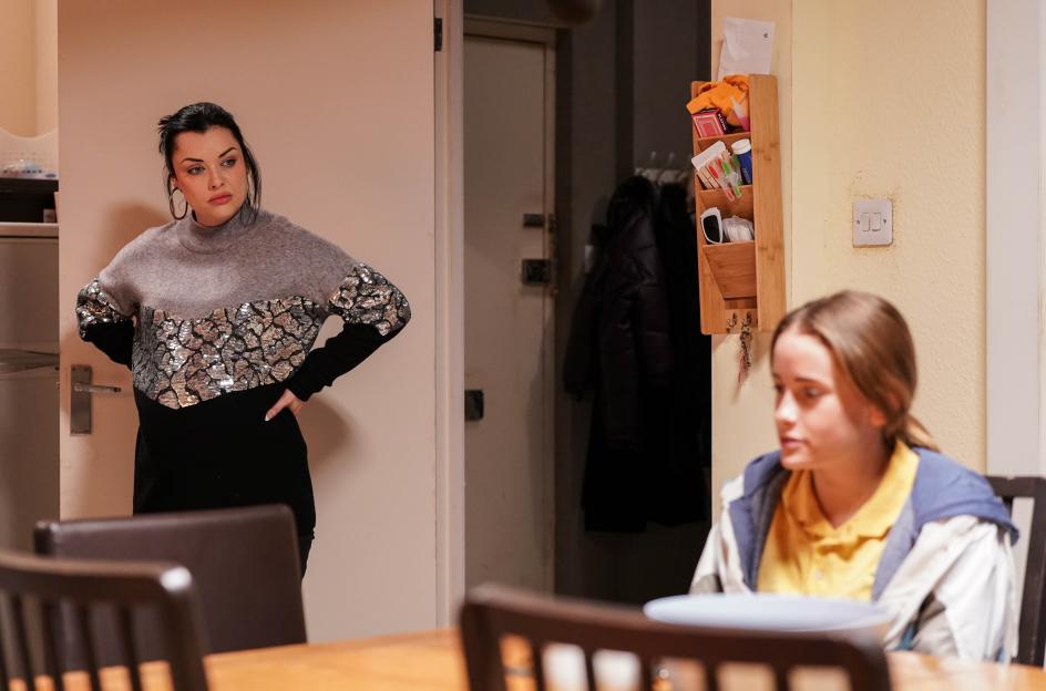 Whitney Dean standing with hands on hips, looking towards Britney, who is seated at a table.