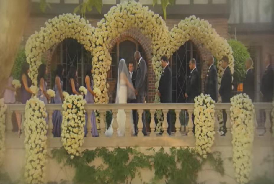 A wedding ceremony with a bride and groom standing before an archway of white flowers, with bridesmaids and groomsmen on either side, all on a balcony adorned with flowers.