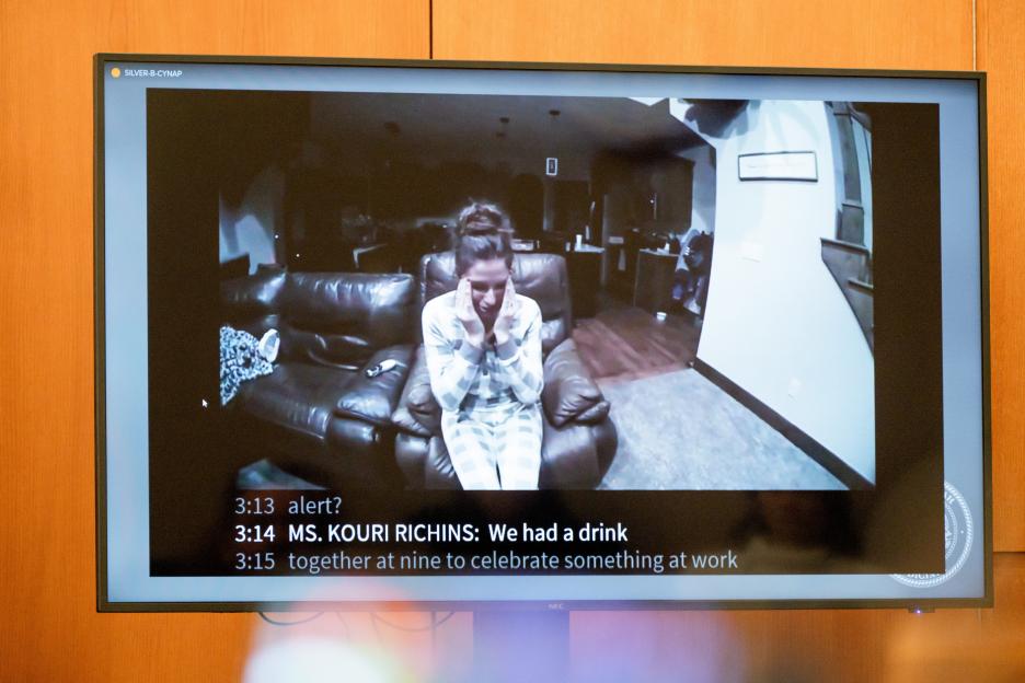 Kouri Richins is seen on a courtroom monitor in surveillance footage, wearing pajamas and sitting on a couch with her hands to her head, with a transcript of her testimony shown below.