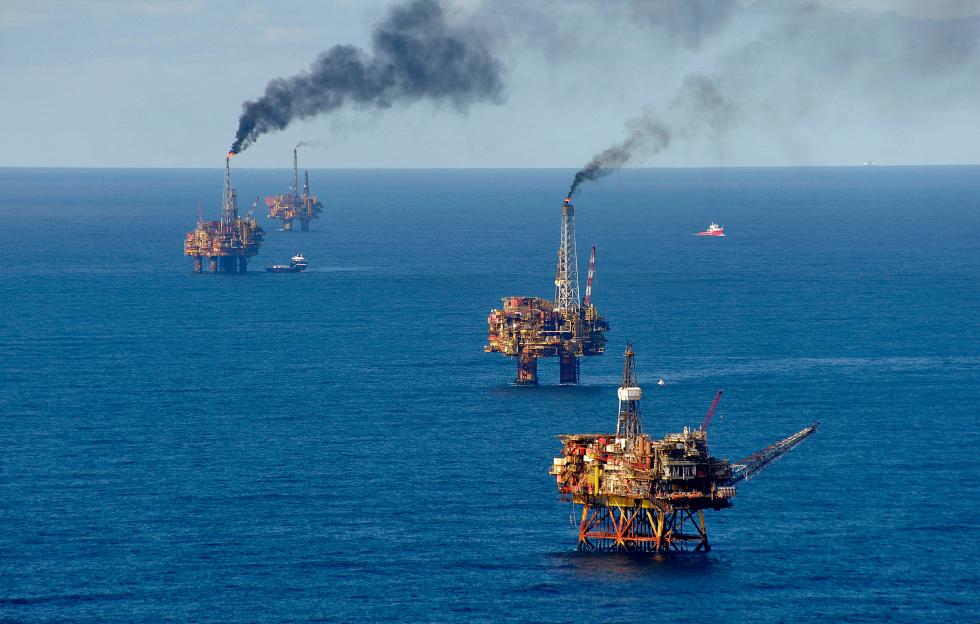 Aerial view of several oil production platforms in the North Sea, with plumes of black smoke rising from some of them.