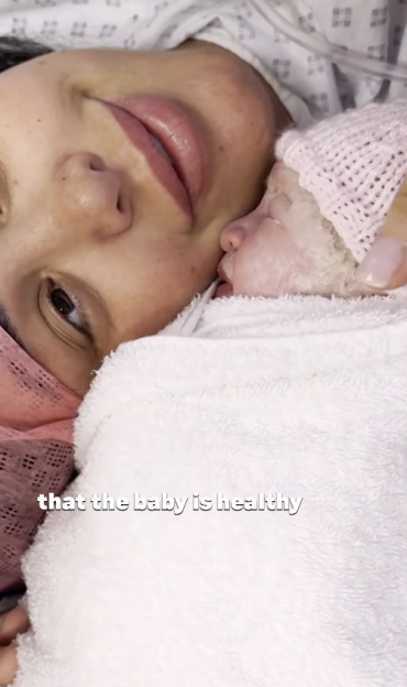 A newborn baby in a pink knit hat, wrapped in a white blanket, resting on its mother's chest.
