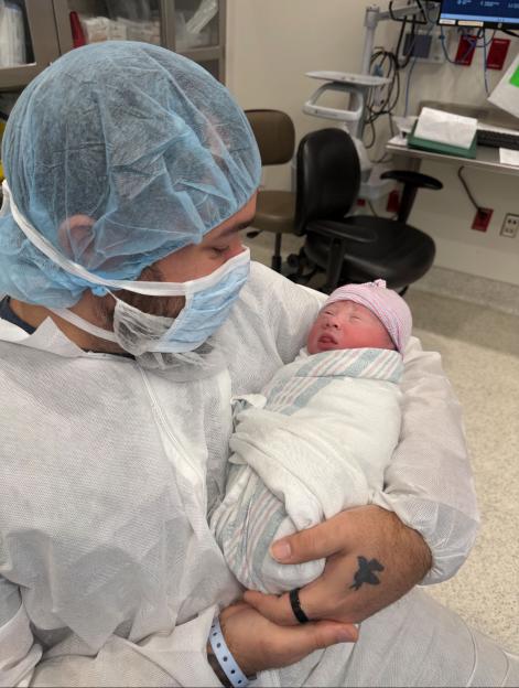 A father in medical scrubs, including a face mask and hairnet, holding his newborn baby in a hospital room.