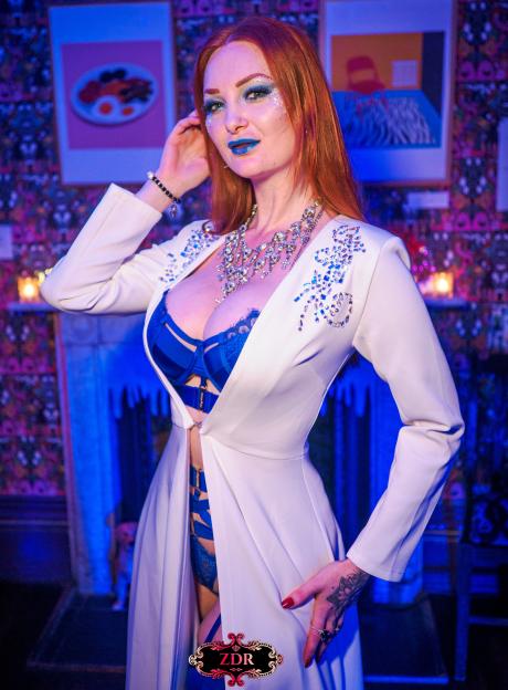 Zara Du Rose, the event organizer, in an open white coat, blue lingerie, and glittery makeup.