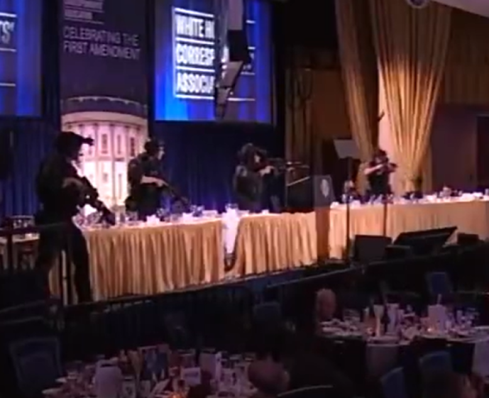 Four armed security guards stand on a stage in front of a banner for the White House Correspondents Association.