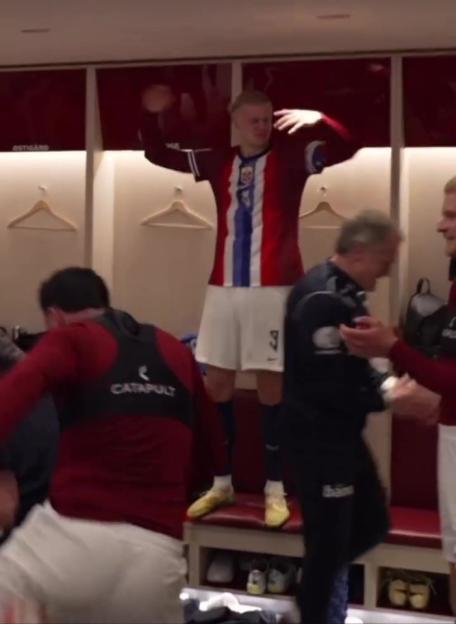 Erling Haaland stands on a bench in a locker room, arms raised, celebrating with teammates.