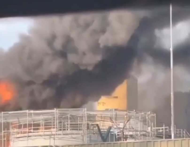 Thick black smoke and orange flames rise from a burning industrial facility.