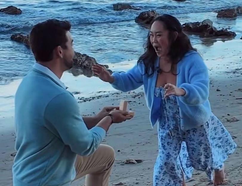 A man on one knee proposing to a woman on a beach, who reacts with an open mouth and outstretched arms.