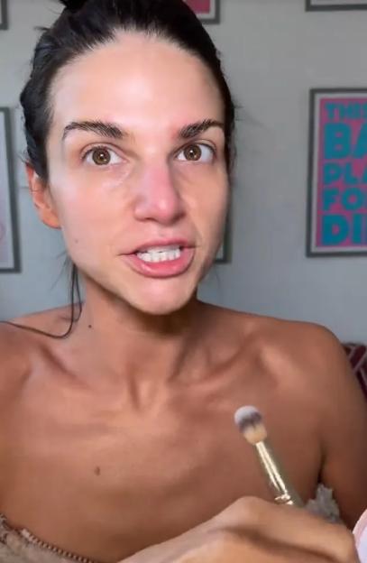 A woman with her hair pulled back, holding a makeup brush, speaks directly into the camera.