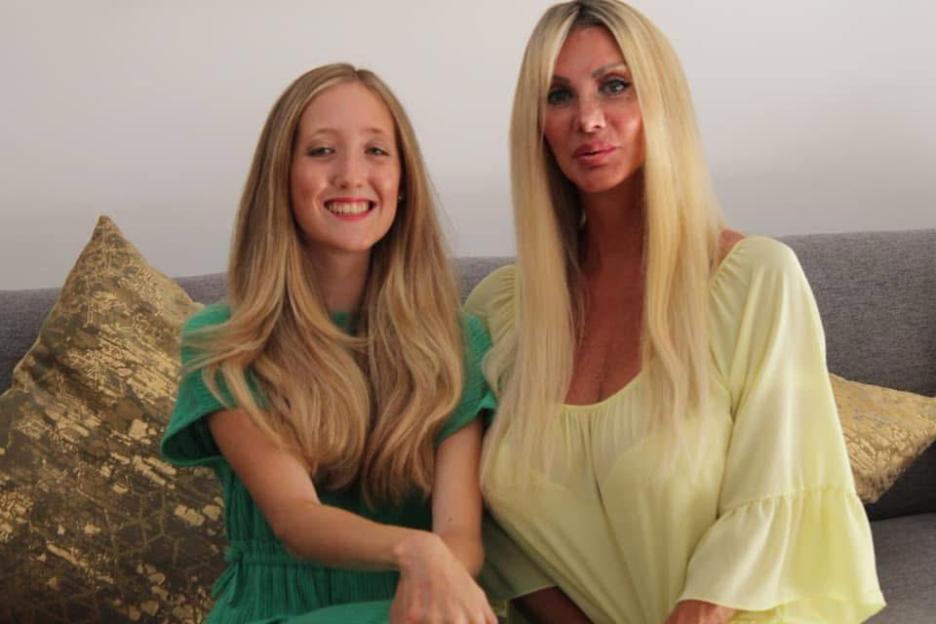 Two women, Sarah Burge and another woman, both with long blonde hair, sitting on a gray couch with gold patterned pillows.