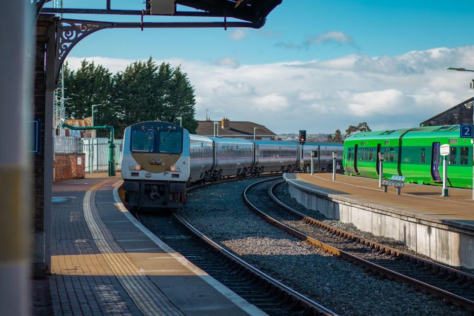 Trains on Drogheda MacBride train station in Ireland, with rail platforms and two trains passing by on a sunny day.