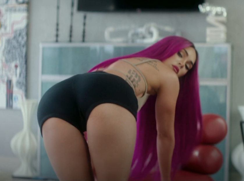 Megan Fox with long pink hair and tattoos, leaning forward in black shorts.