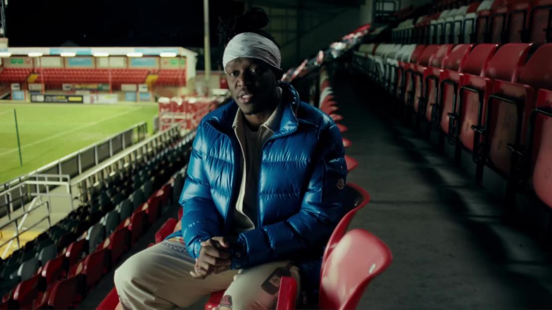 KSI seated in an empty stadium wearing a blue puffer jacket and white bandana.
