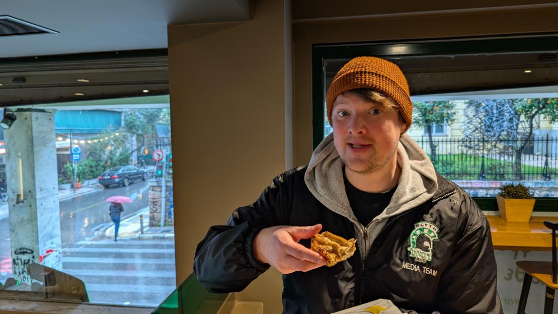 Man holding a pastry in a bakery with a rainy street scene outside the window.