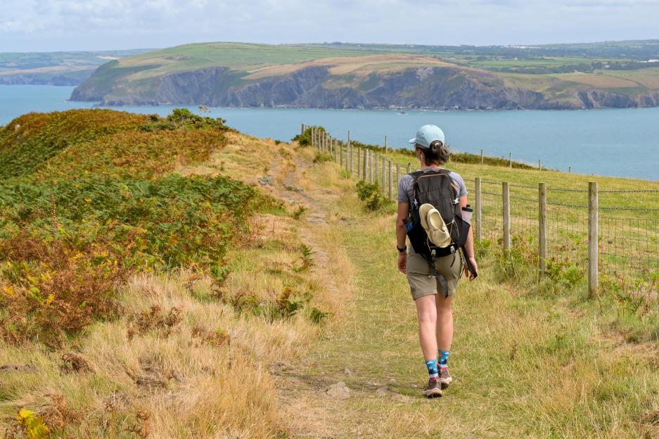 A person walks along the Wales Coast Path near Dinas Head with the ocean and distant coastline in view.