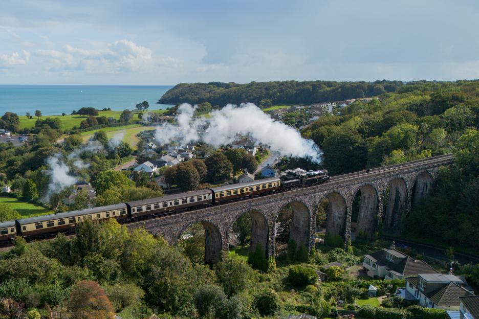Aerial view of the Dartmouth Steam Train on Hookhills Viaduct in Devon, England, with the sea and coastline in the background.