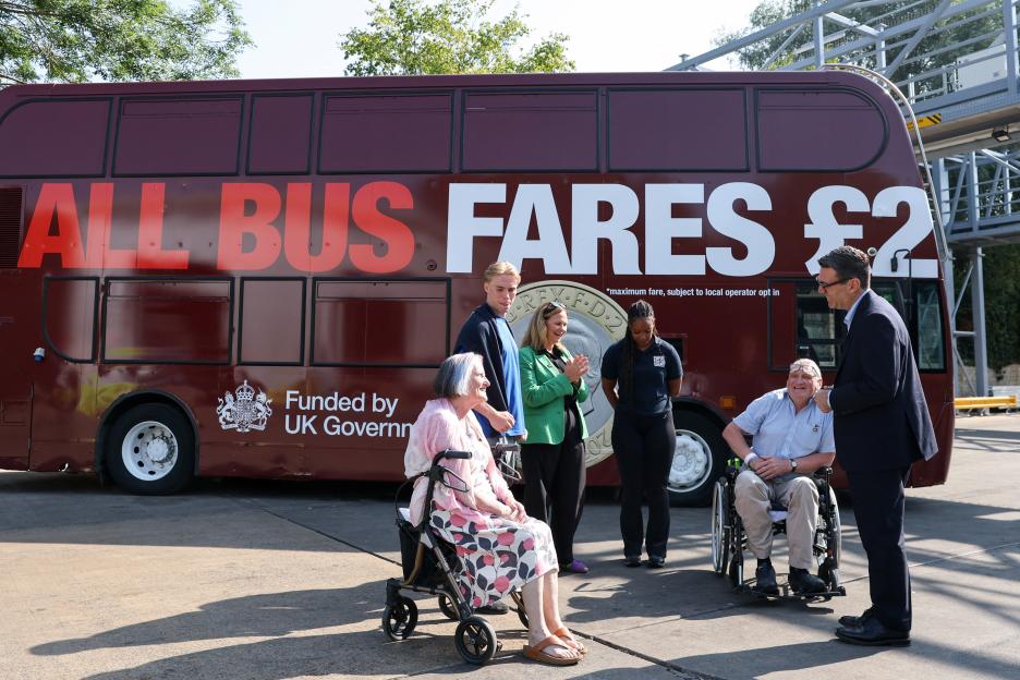 Prime Minister Andy Burnham and Mayor Helen Godwin speak with local bus users, including two in wheelchairs, in front of a bus advertising "ALL BUS FARES £2" funded by the UK Government.