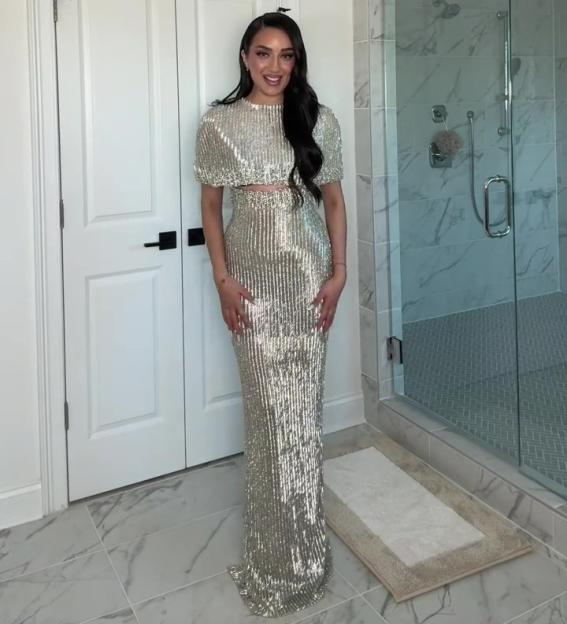 A woman wearing a shiny, silver, two-piece sequined dress, consisting of a short-sleeved crop top and a floor-length skirt.