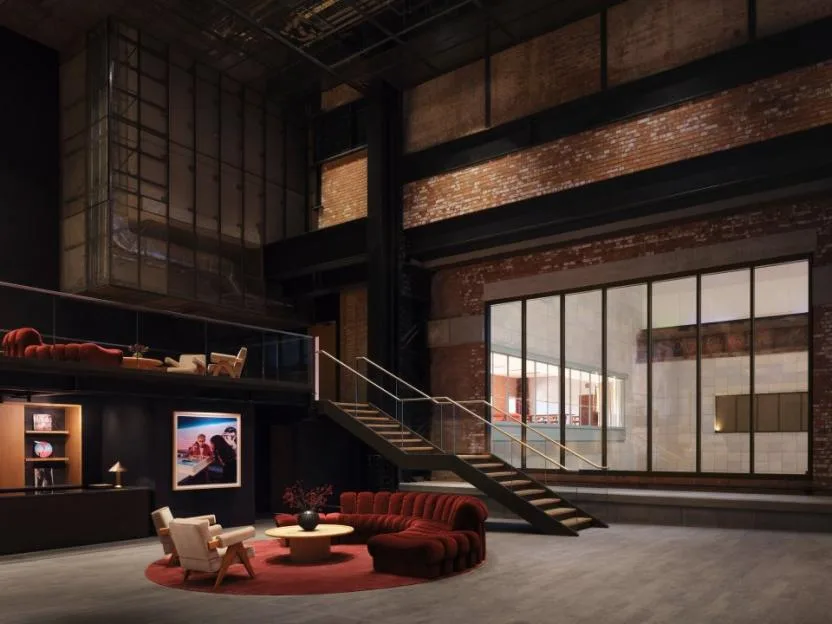 Interior of the Apple Music Hall foyer with a red velvet sofa, lounge chairs, a wooden coffee table on a round red rug, and a modern staircase leading to an upper level.