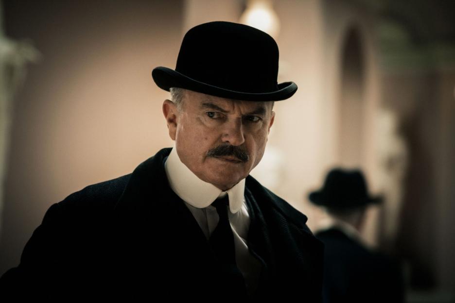 Sam Neill as Inspector Campbell in Peaky Blinders, wearing a black bowler hat and coat, with a stern expression.
