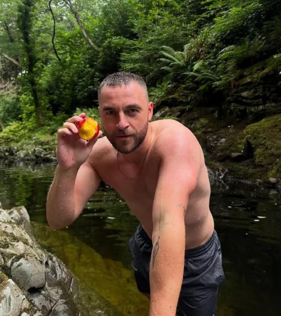 A man holds a half-eaten peach while standing in a body of water, surrounded by lush green foliage.