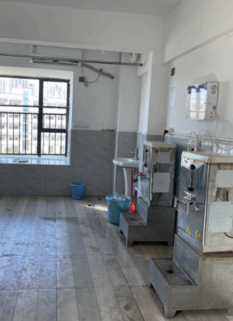 A room with two water dispensers, a sink, and two blue buckets, with a barred window on the left.