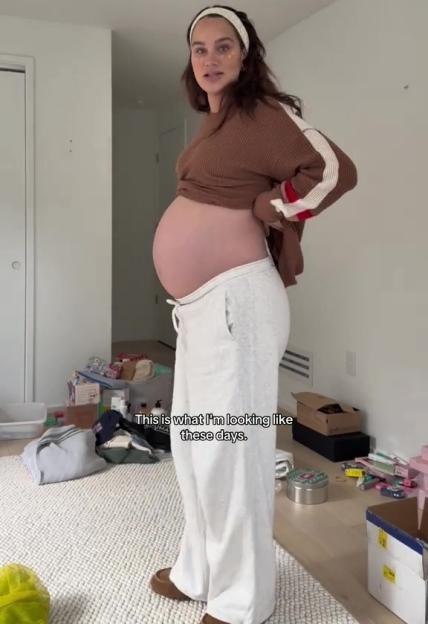 Pregnant woman standing sideways in a room, revealing her bare baby bump, with the text "This is what I'm looking like these days."