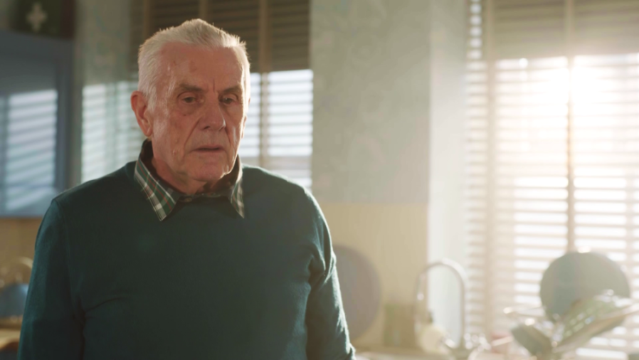 An elderly man with white hair, wearing a teal sweater over a plaid shirt, looks forward with a concerned expression.