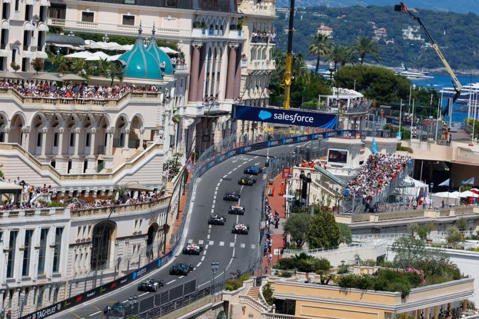 A wide shot of Formula 1 race cars on a street track through a city, with spectators on balconies and grandstands.