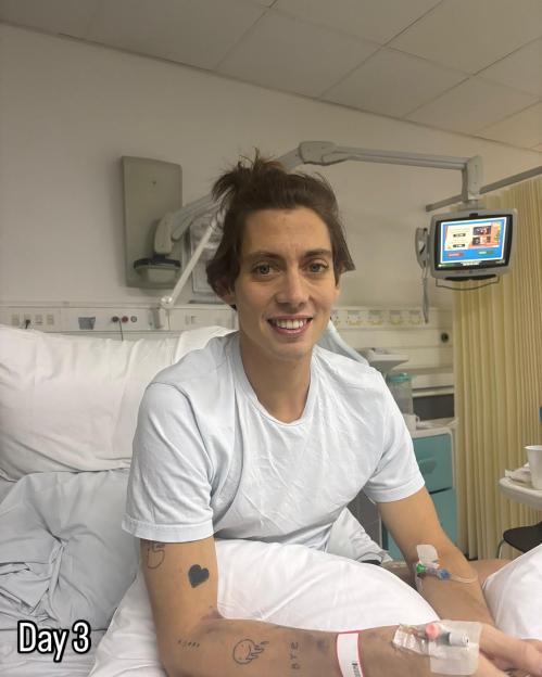 A young man with an IV in his arm and small tattoos, including a black heart, on his arm, smiles while sitting up in a hospital bed.