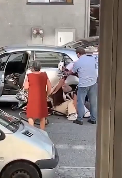 Several people help a fallen cyclist next to two cars.
