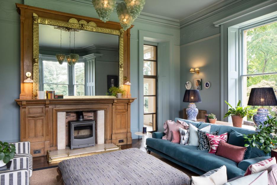 A living room with light blue walls, a large wooden fireplace, a blue velvet sofa, and an ornate mirror.