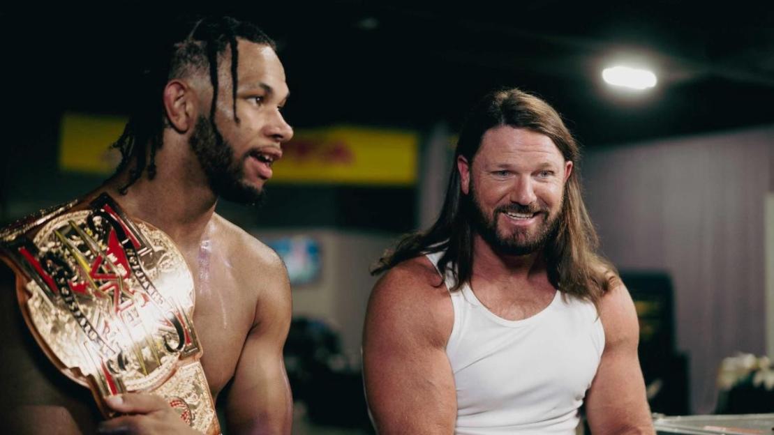 Two male wrestlers backstage, one with a championship belt, smiling and talking.