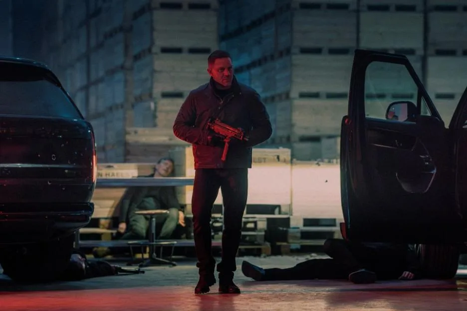 Tom Hardy as Harry Da Souza holding a submachine gun, standing between two cars with bodies on the ground.