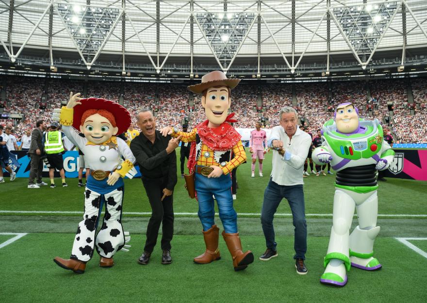 Tom Hanks, Woody, Tim Allen, Jessie, and Buzz Lightyear pose on a soccer field in a stadium full of people.