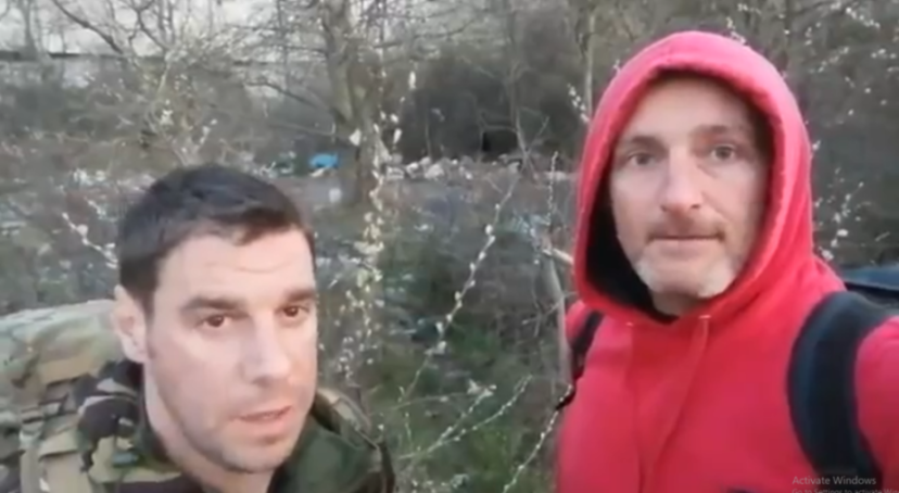 Two men, one in a camouflage jacket and the other in a red hoodie, stand outside among bare trees.