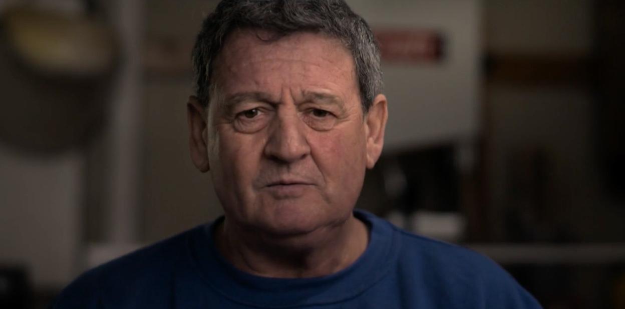 A middle-aged man with dark gray hair and a blue sweatshirt looks into the camera with a neutral expression.
