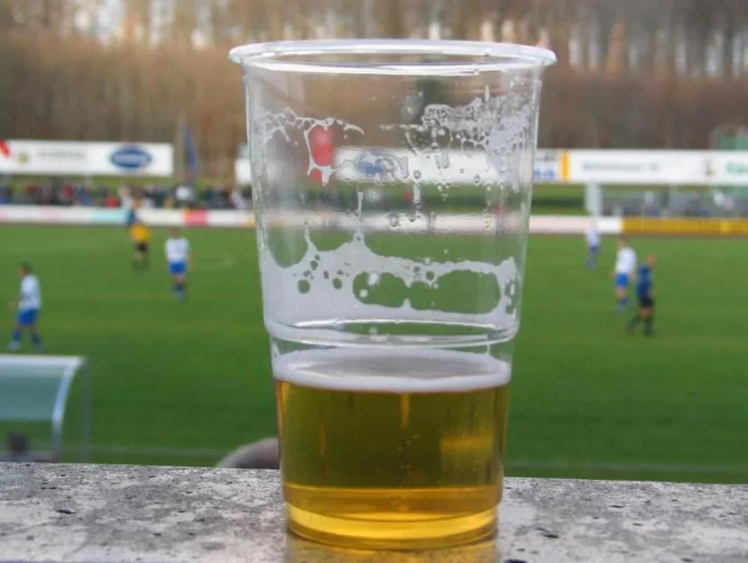 A plastic cup of beer sits on a wall overlooking a soccer field with players.
