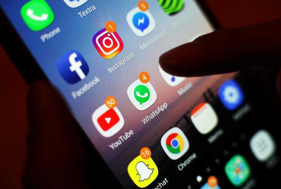A mobile phone screen displays icons for social media apps including Facebook, Instagram, YouTube, and WhatsApp, with notifications on Instagram, YouTube, and WhatsApp.
