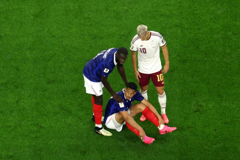 William Saliba sitting on the pitch after an injury, flanked by Dayot Upamecano and Dani Olmo.